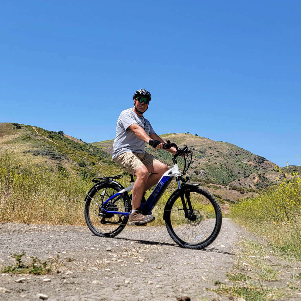 Person riding an E-Joe electric bike on a scenic trail surrounded by green hills under a clear blue sky.