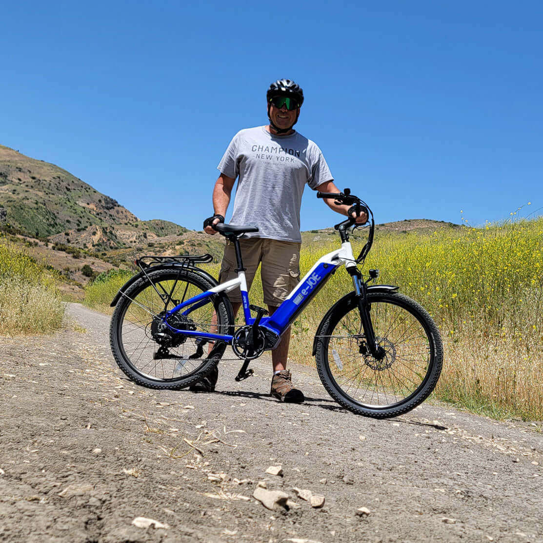 Man in a gray t-shirt standing next to a blue electric bike on a dirt path surrounded by greenery.