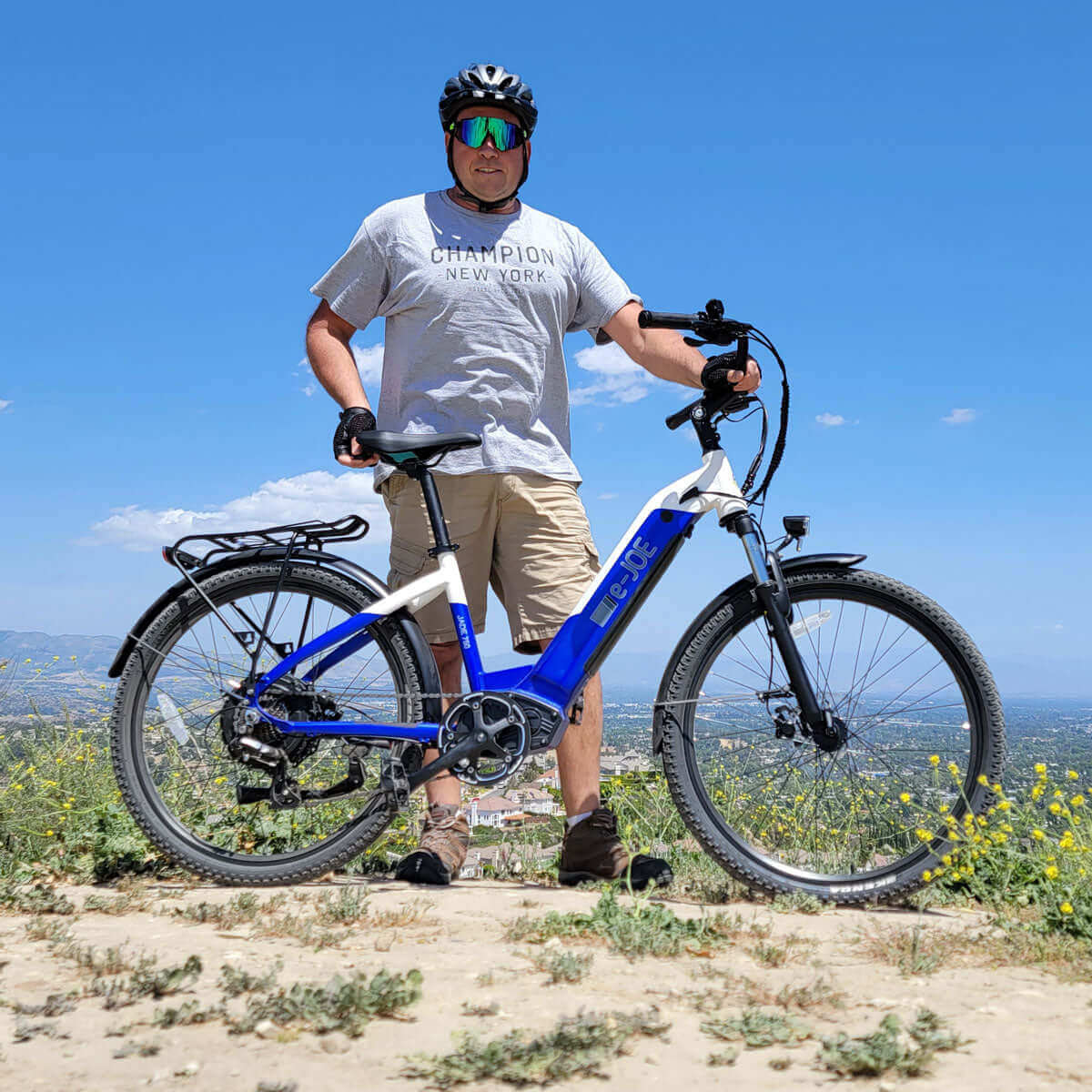 Man wearing sunglasses and a Champion shirt standing next to an E-Joe electric bike on a sunny day outdoors.