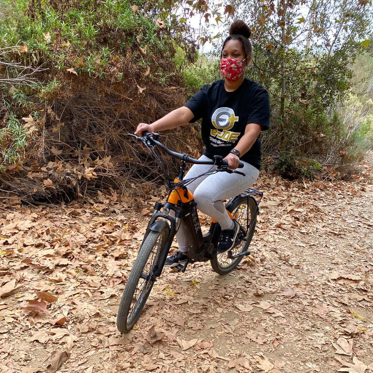 Woman riding an electric bike on a wooded trail, wearing a black t-shirt and mask, surrounded by autumn leaves.