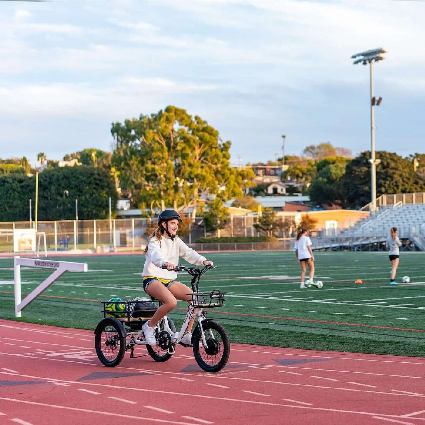 A young woman riding a step-thru electric trike on a track field with trees and stadium in the background.
