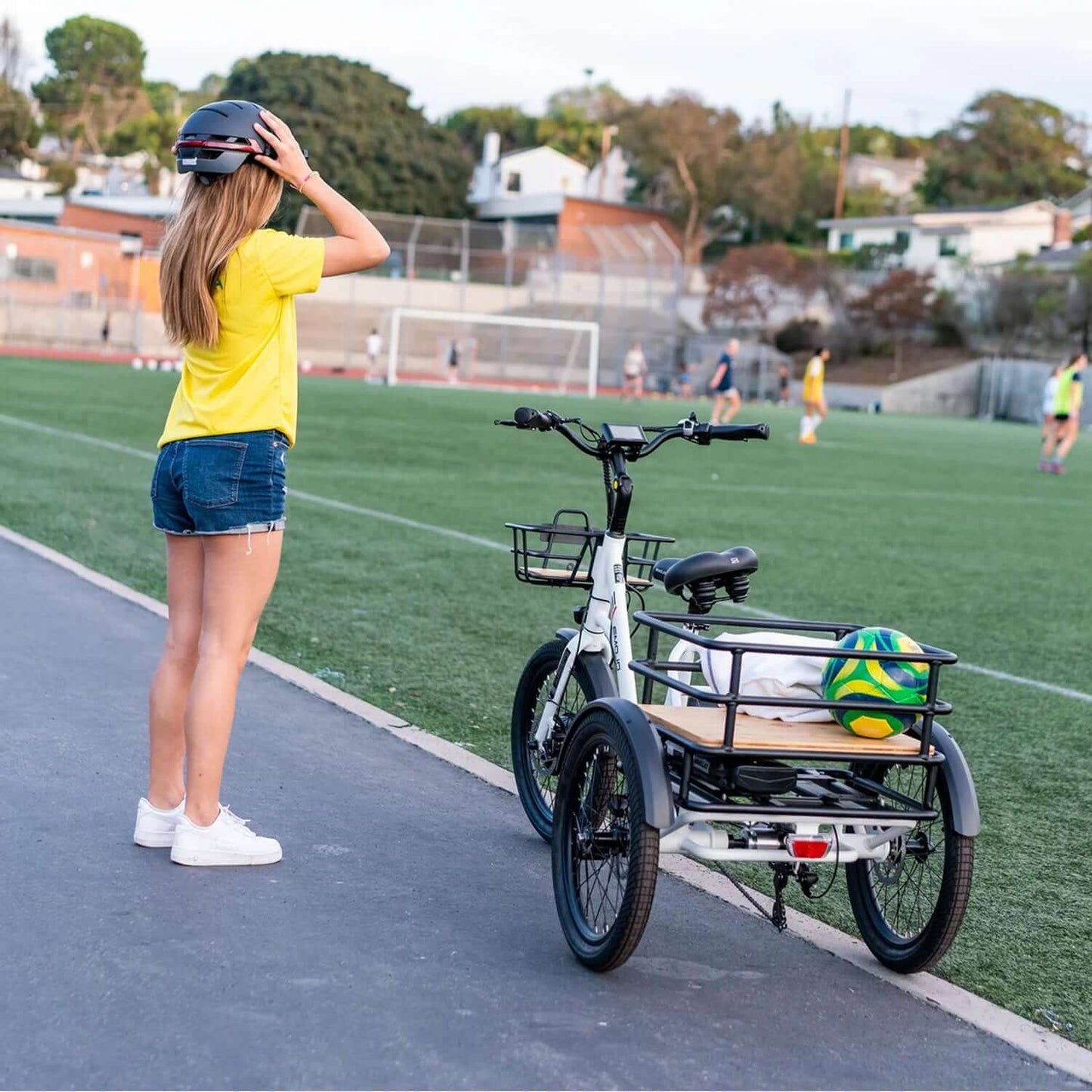 A woman wearing a helmet stands by an Emojo Bison S electric trike on a sports field with a soccer ball.