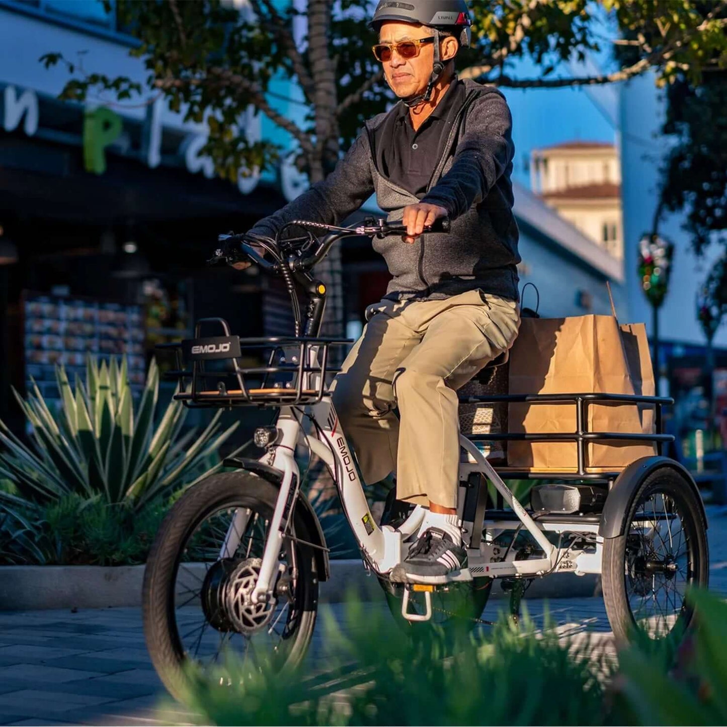 Man riding Emojo Bison S 48V electric trike with shopping bags in sunny urban environment.