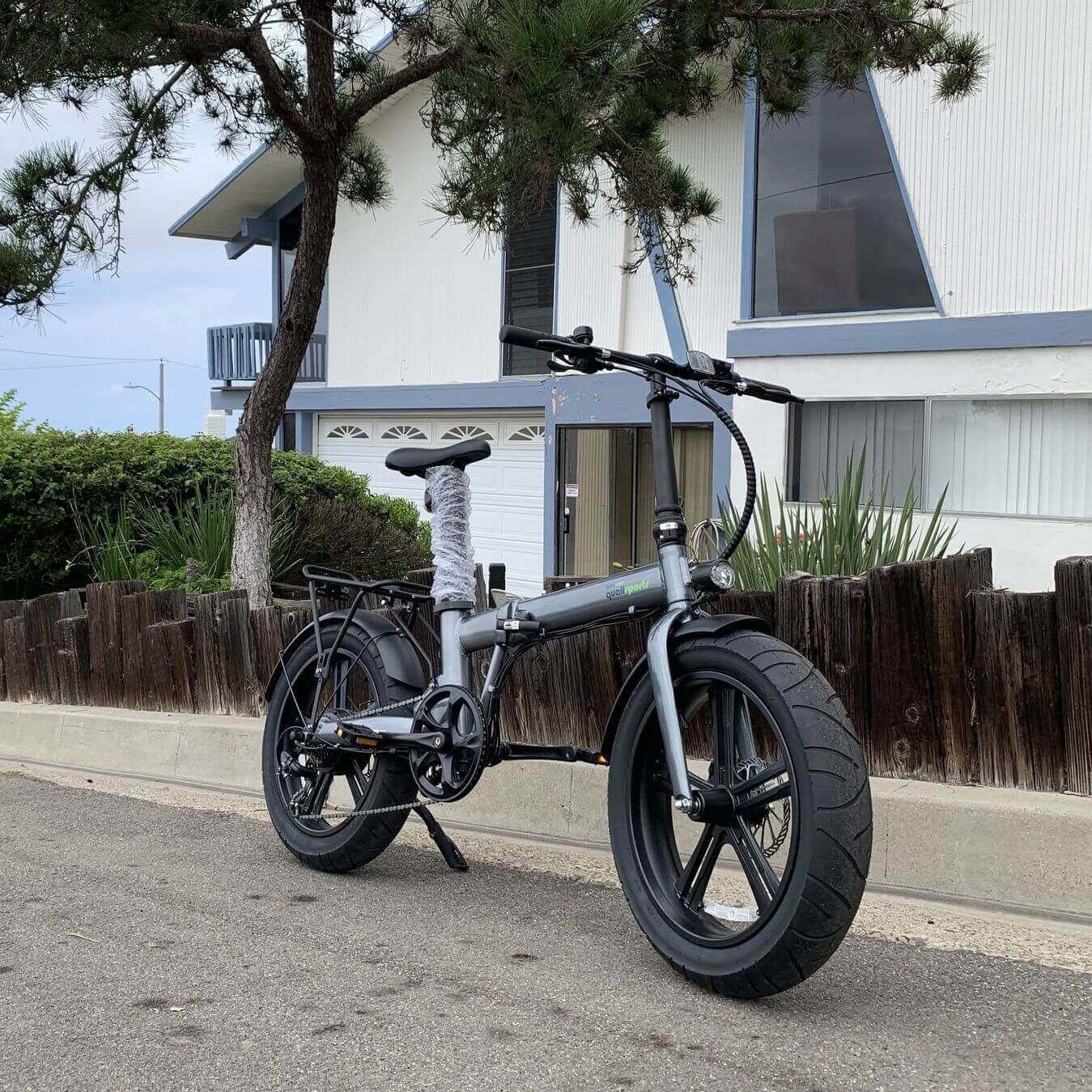 Qualisports Beluga STD/PLUS fat tire folding electric bike parked on a street with greenery in the background.