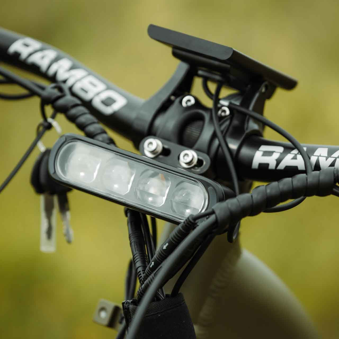 Bicycle handlebar with 'RAMBO' branding and a light attachment against a blurred natural background.