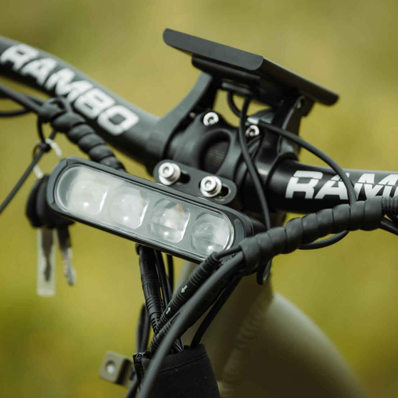 Bicycle handlebar with 'RAMBO' branding and a light attachment against a blurred natural background.