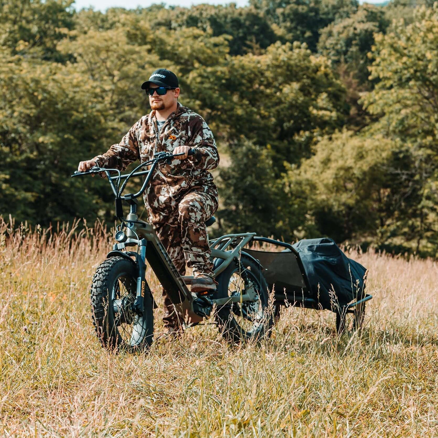 Person in camouflage gear riding a four-wheeled vehicle with a trailer through a forested area.