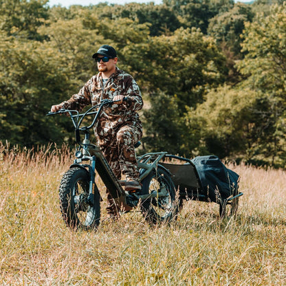 Person in camouflage gear riding a four-wheeled vehicle with a trailer through a forested area.