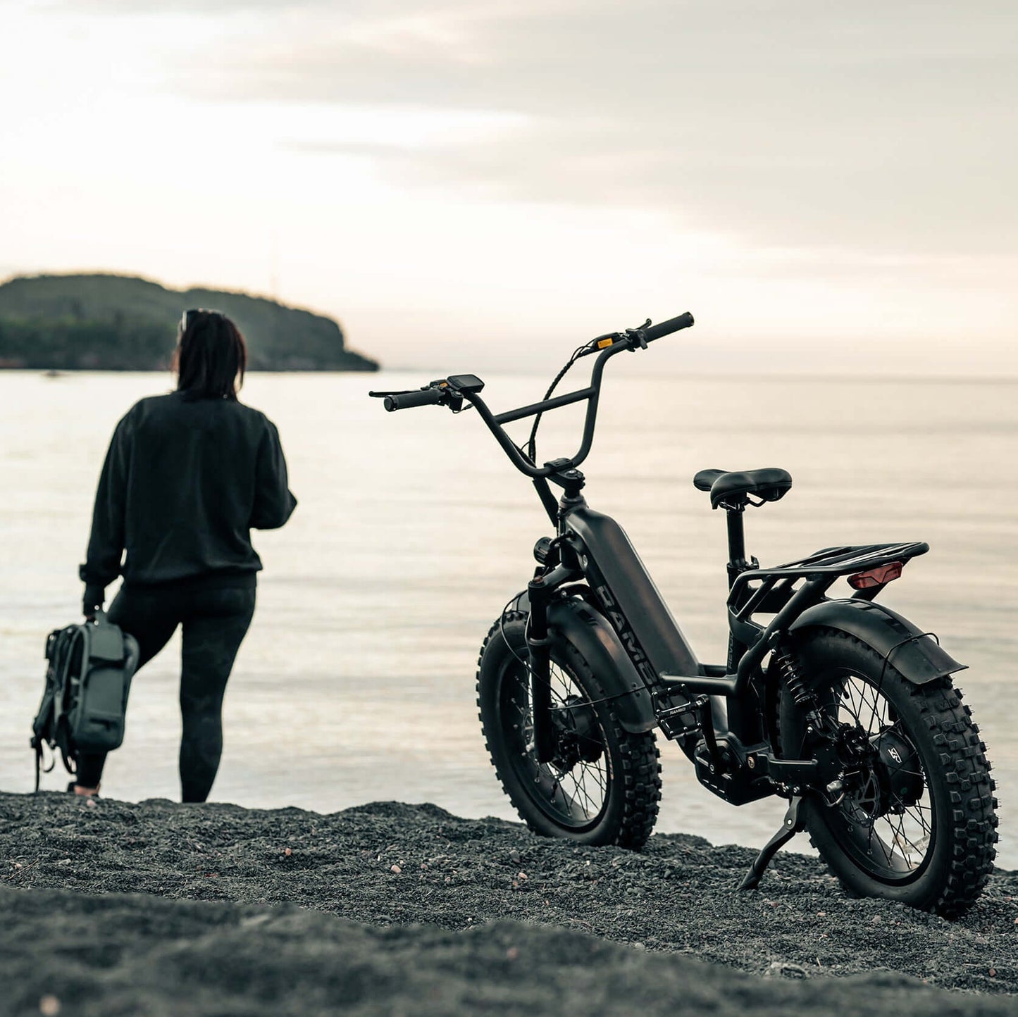 Person with a suitcase standing next to an electric bike on a beach at sunset.